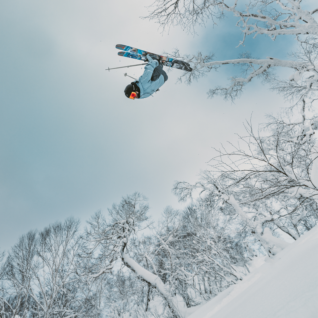 A skier mid-air performing a trick on snowboard and skis, surrounded by snow-covered trees under a pale winter sky, wearing VALLON Crossroads ski goggles