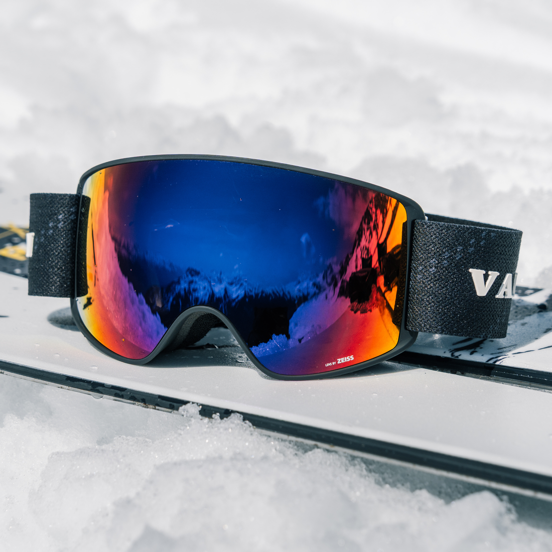 Black-Purple Crossroads ski goggles from VALLON with a mirrored blue-to-orange reflective lens, resting on a pair of skis in the snow.