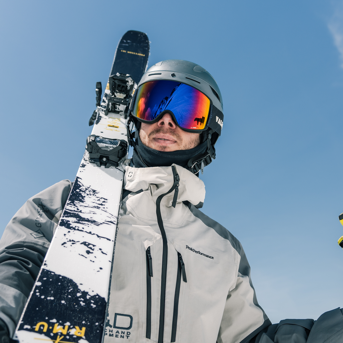 A close-up shot of a skier wearing a silver helmet, Crossroads ski goggles from VALLON with purple lenses, and a light gray ski jacket, holding a ski up against a clear blue sky. The ski features a white and black snow pattern with yellow branding.