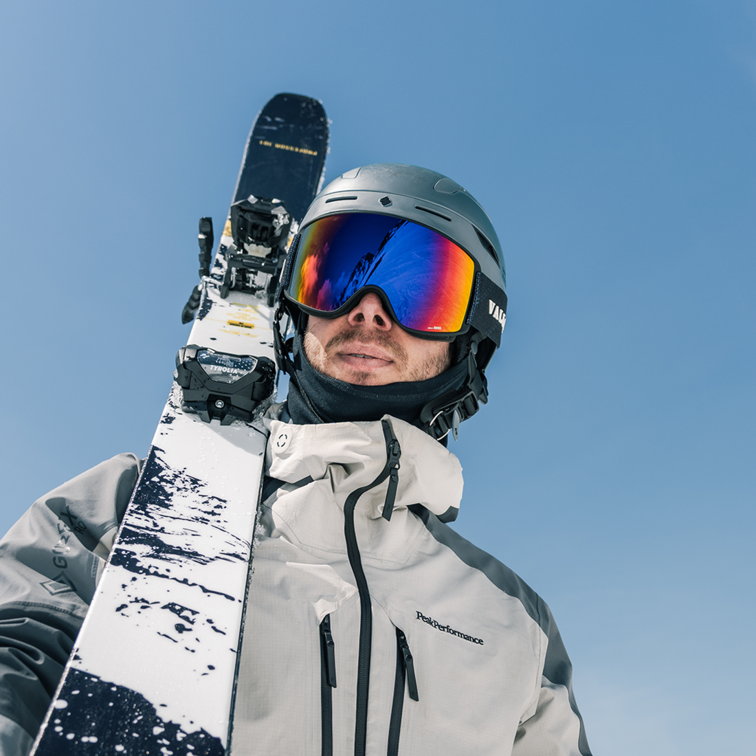 A close-up shot of a skier wearing a silver helmet, Crossroads ski goggles from VALLON with purple lenses, and a light gray ski jacket, holding a ski up against a clear blue sky. The ski features a white and black snow pattern with yellow branding.