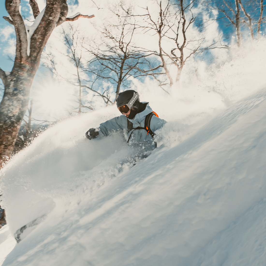 A skier carving through deep powder snow on a steep slope, surrounded by bare winter trees and bright blue sky. The skier is wearing a light blue ski jacket, VALLON Crossroads ski goggles, black helmet, and orange-accented harness.
