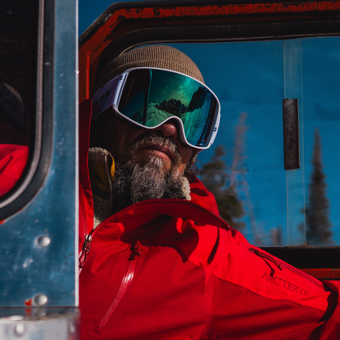 White-Green |A close-up lifestyle shot of a man wearing White-Green VALLON Crossroads ski goggles, a red Arc'teryx ski jacket and a brown beanie, sitting inside a vehicle with blue-tinted windows.