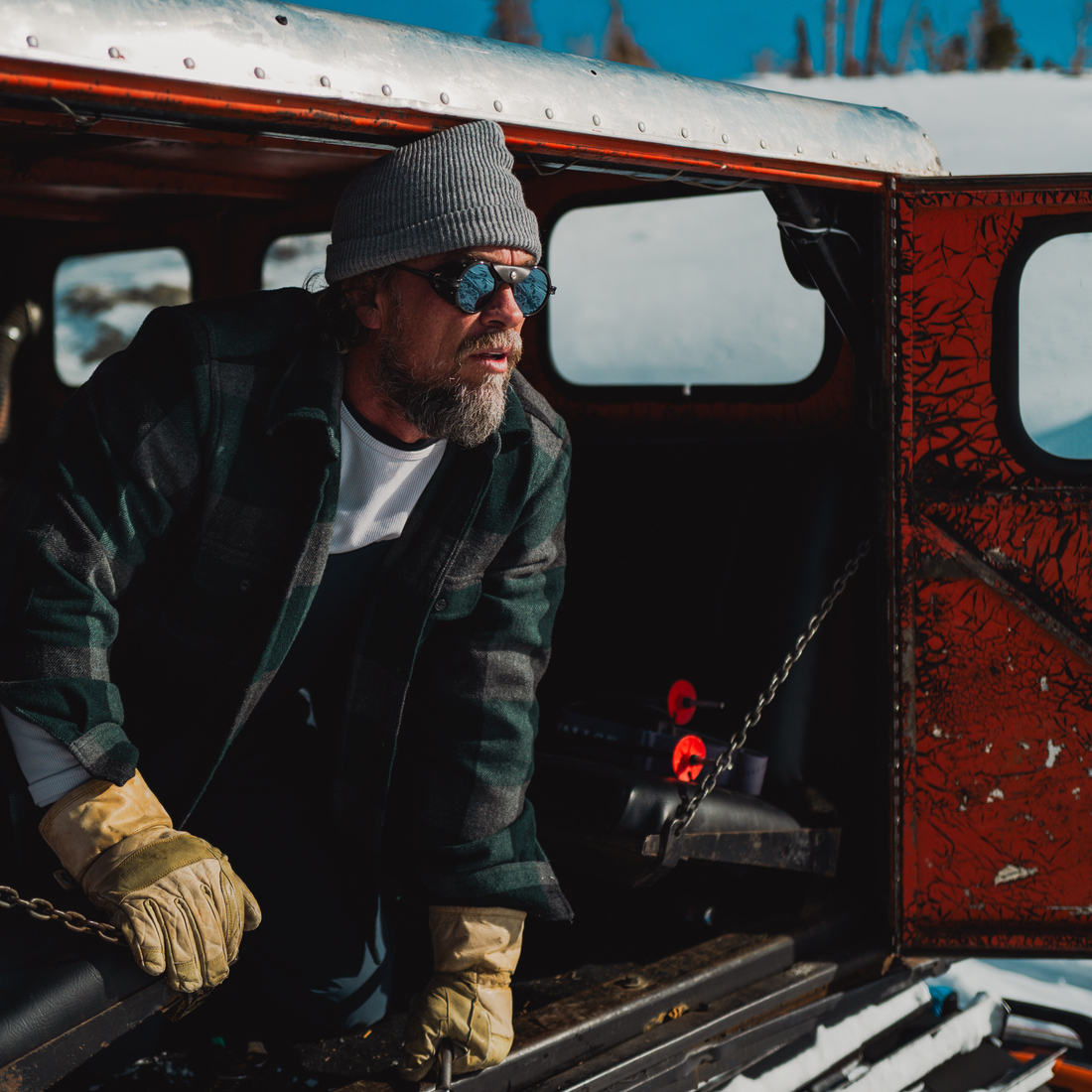 A man wearing a gray knit beanie, Brown Heron Glacier sunglasses from VALLON, a dark green plaid flannel jacket over a white t-shirt, and tan work gloves leans out of the open back of a rustic red and silver truck in a snowy outdoor setting.