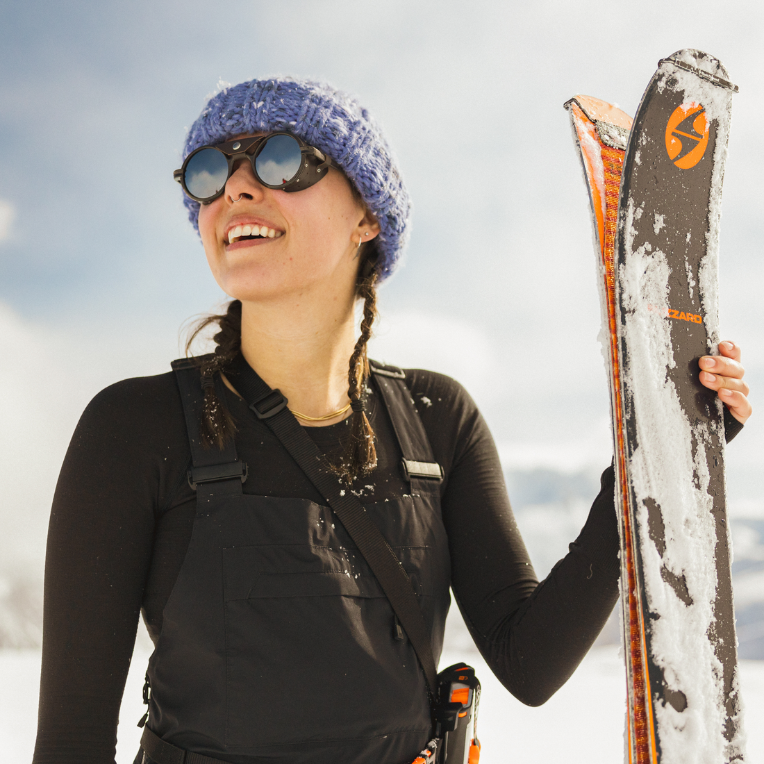 A woman wearing a blue knit beanie, Brown Heron Glacier sunglasses, and black winter overalls holds a pair of skis with snow on them against a snowy outdoor background.