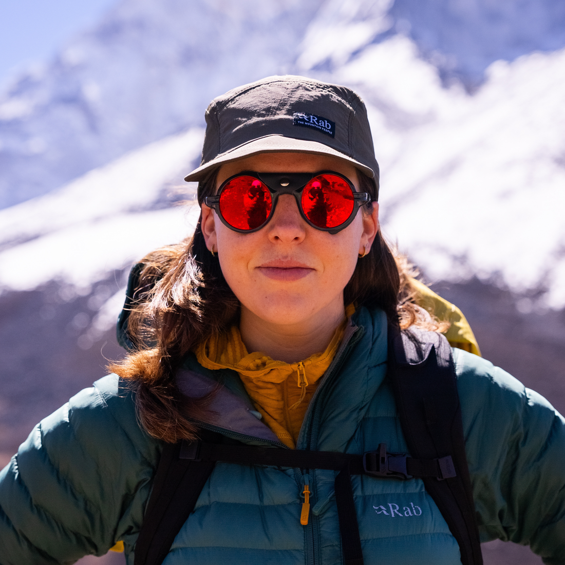 Adriana Brownlee stands outdoors in front of snowy mountains, wearing a dark gray Rab cap, VALLON Heron Glacier glasses with red lenses, a teal Rab puffer jacket, and a yellow inner layer.