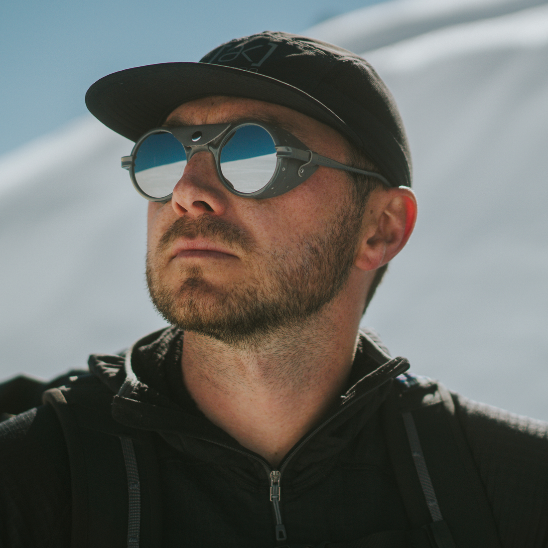 A close-up portrait of a man wearing round Cat. 4 Heron Glacier sunglasses with gray frames, a black baseball cap, and a black zip-up jacket against a bright outdoor background.