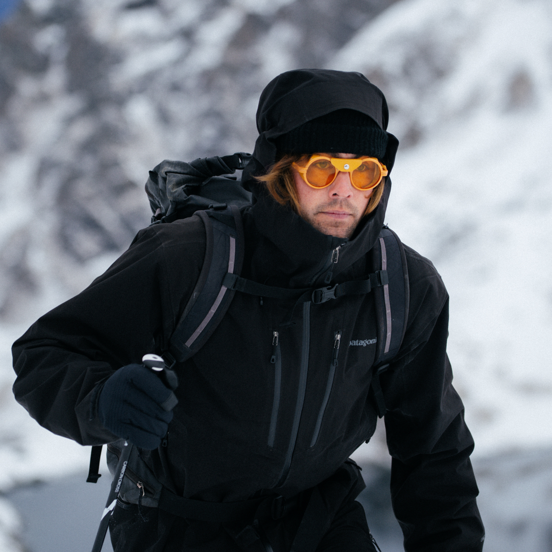 A man in a snowy mountain setting wearing a black Patagonia jacket, black beanie, black gloves, and Low-Light Heron Glacier sunglasses from VALLON.
