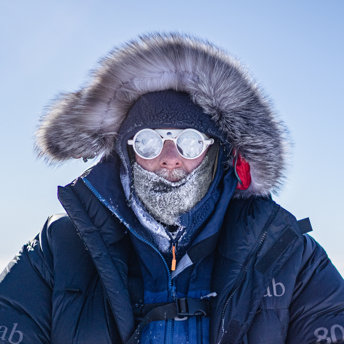 A close-up portrait of a person outdoors in extreme cold, wearing a dark insulated parka with a large gray fur-trimmed hood, a dark knit beanie, round white-framed, mirror-coated Heron Glacier sunglasses from VALLON, and a frosty face covering against a pale blue sky.