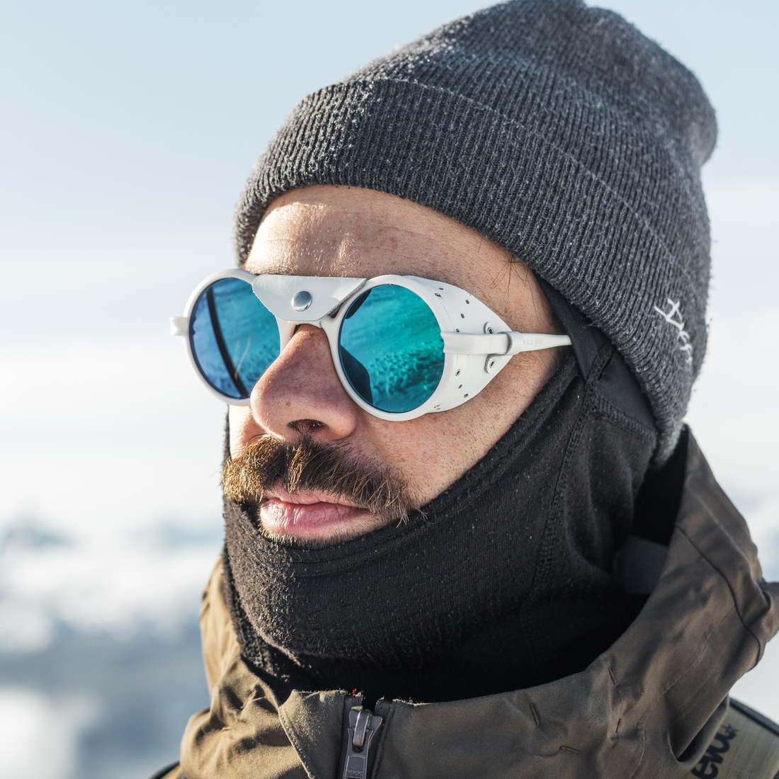 A close-up of a man wearing white Heron Glacier sunglasses with blue-green mirrored lenses, a dark gray knit beanie, and a black face balaclava in a snowy outdoor setting.