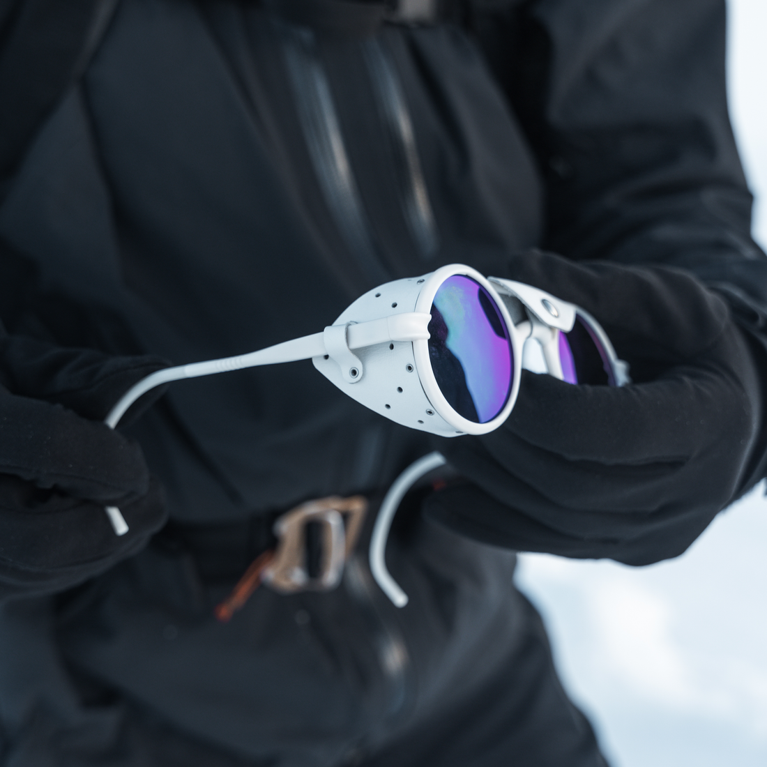 A close-up of a person wearing black winter clothing and gloves, holding a pair of Off-White-Green Heron Glacier ski sunglasses from VALLON with purple mirrored lenses.