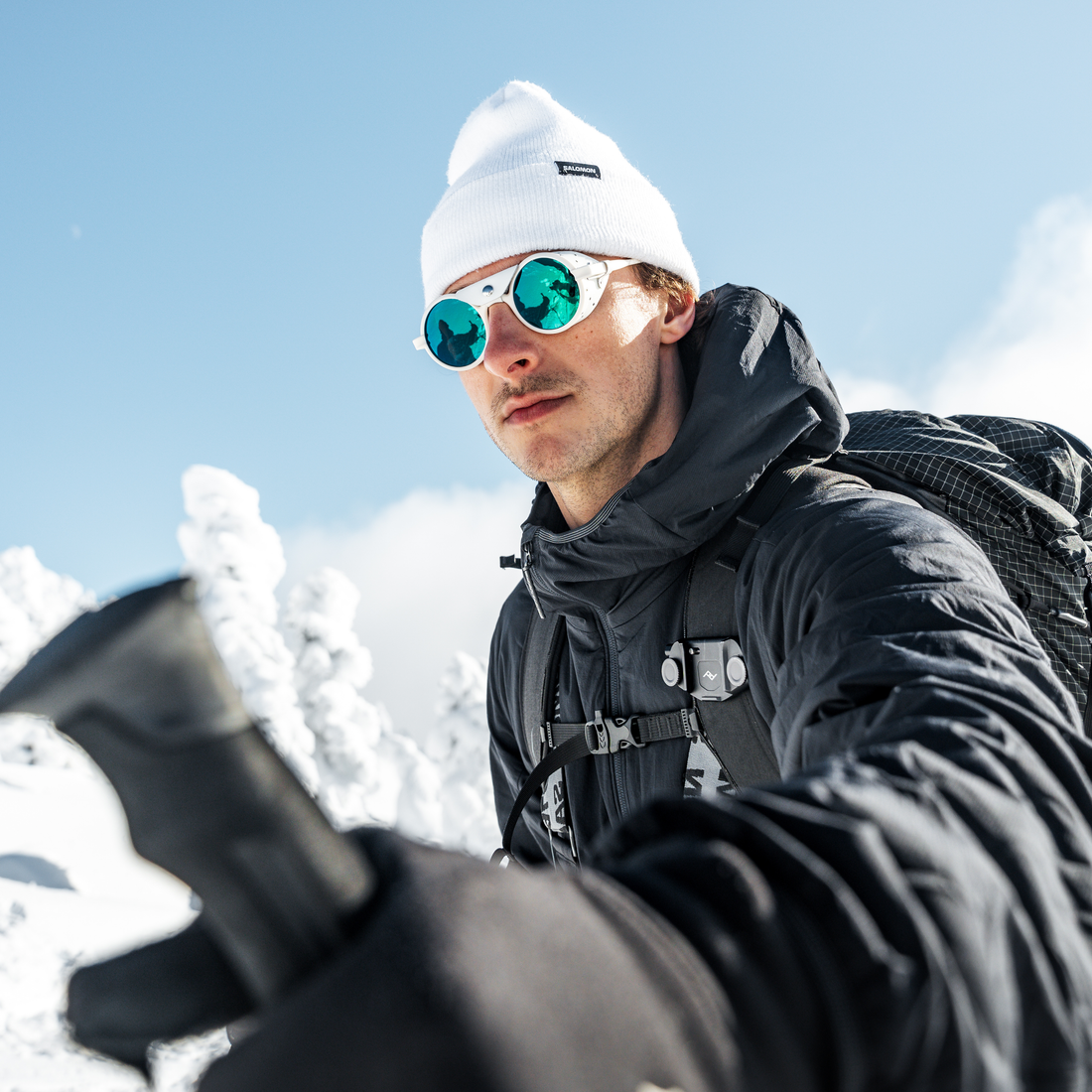 A man wearing a white beanie, white-framed Heron Glacier sunglasses with green mirrored lenses, a black winter jacket, and a black backpack takes a selfie in a snowy outdoor setting under a clear blue sky.