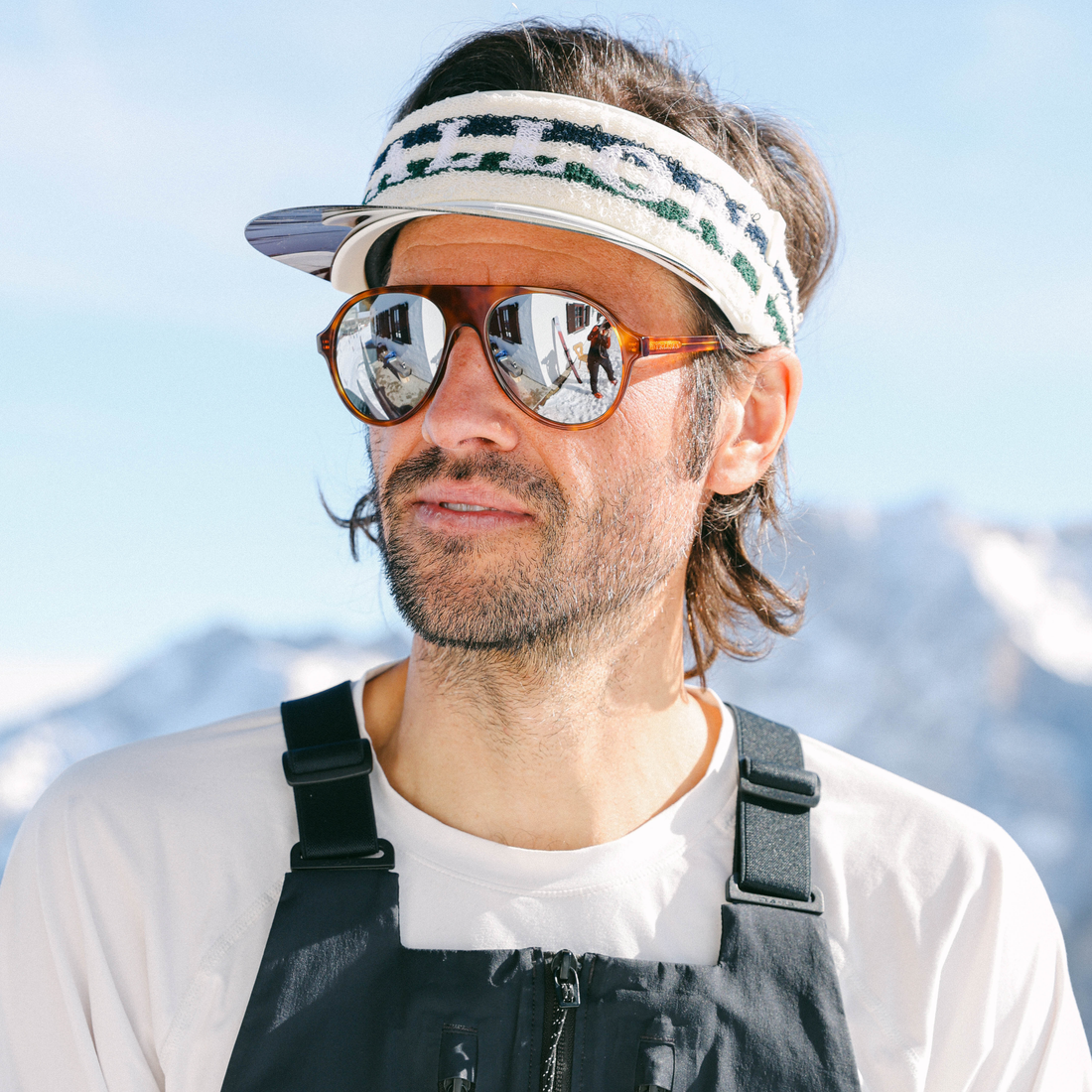 A man outdoors wearing a white VALLON Vizor with green lettering, Tortoise-framed mirrored Hazlewood sunglasses, a white T-shirt, and dark bib overalls, with a blurred mountain background.