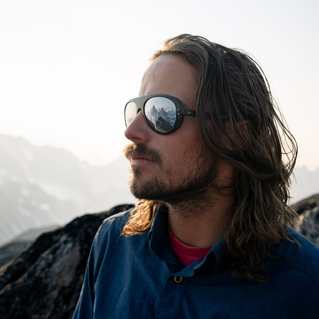 A man with long brown hair and a beard wearing black Ski Aviators sunglasses with a mountain reflection in the mirror lenses, standing outdoors against a soft-focus mountain background.