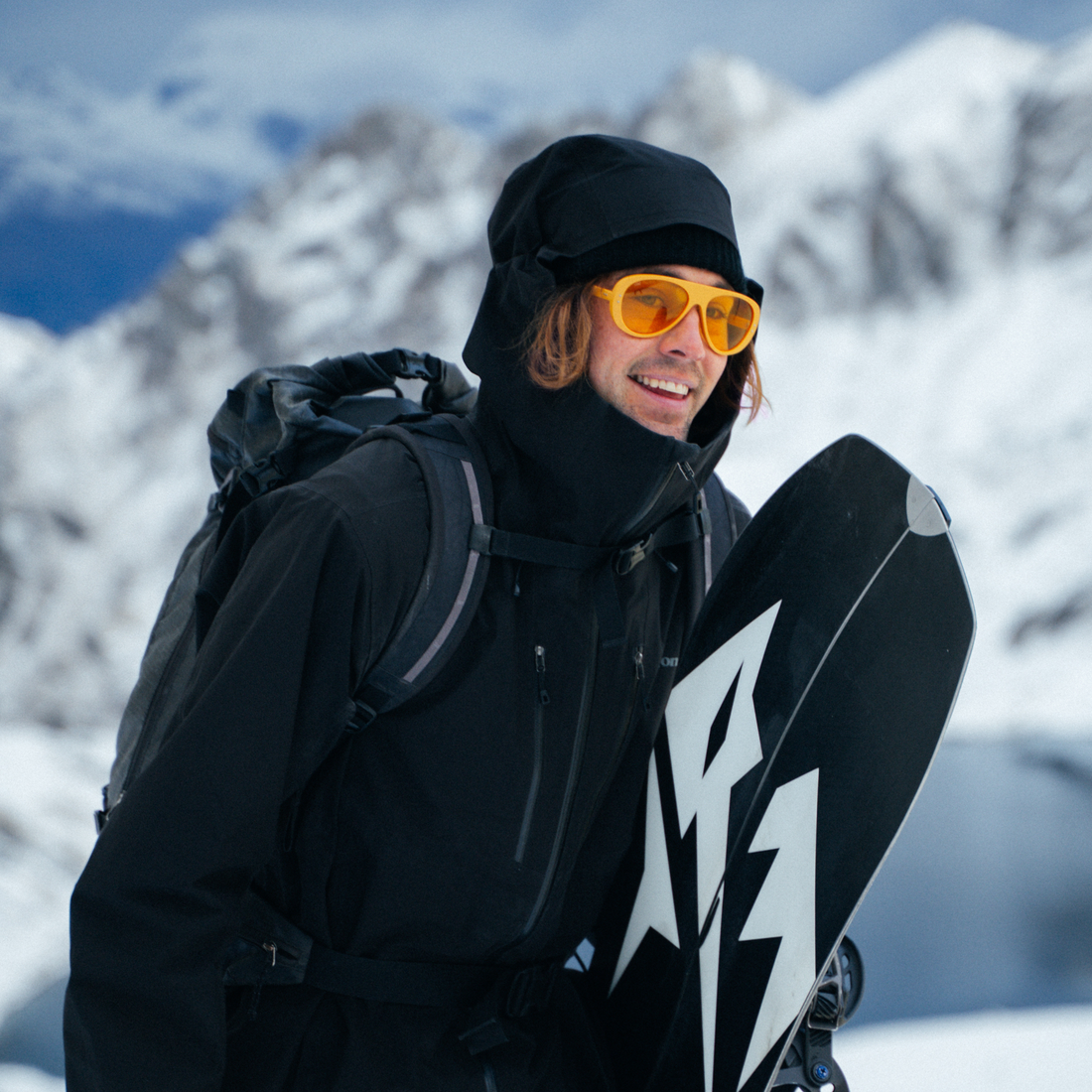 A snowboarder in a black winter jacket, black beanie, and bright yellow-tinted Ski Aviators with low-light lenses holds a black snowboard with a white graphic, set against a snowy mountain background.