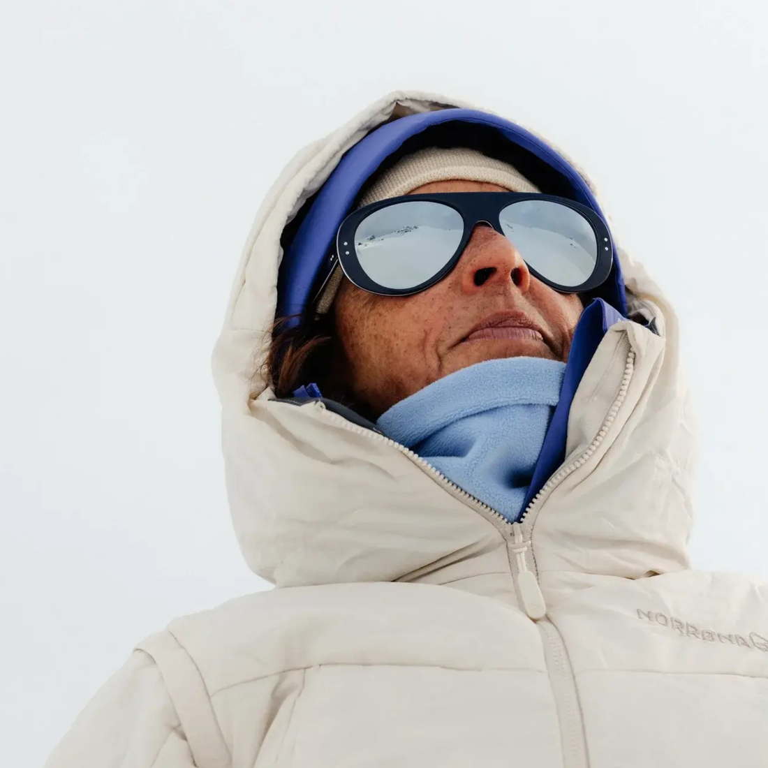 A close-up shot of a person wearing a light beige hooded winter jacket, a light blue neck gaiter, a blue headband, and Tricolor Blue Ski Aviators sunglasses with mirrored lenses from VALLON, set against a bright, overcast white sky.