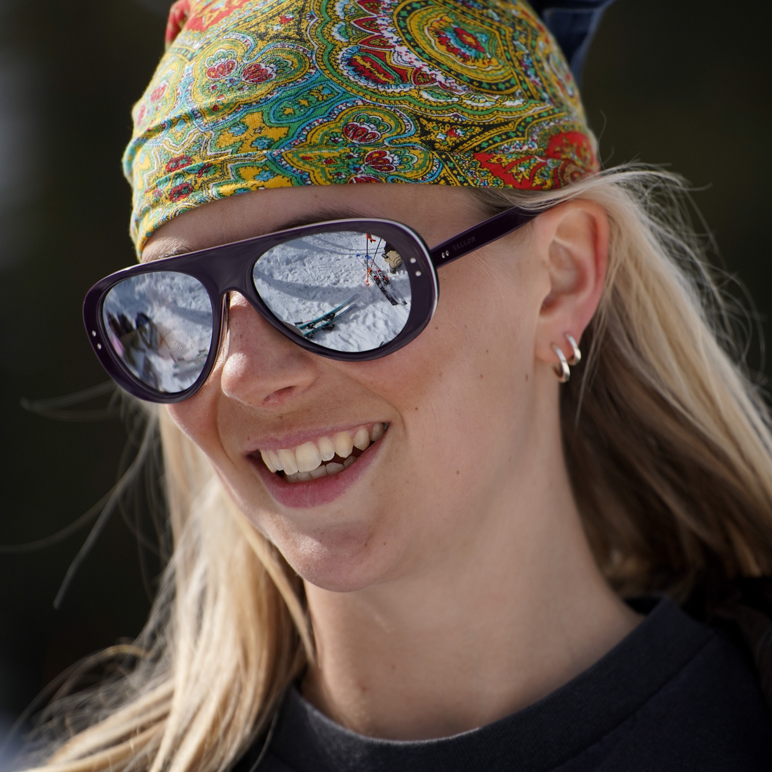 A close-up of a smiling woman wearing purple tricolor Ski Aviators sunglasses from VALLON with a colorful paisley-patterned headband and a black top. The sunglasses have dark frames and reflective lenses showing a snowy outdoor scene.