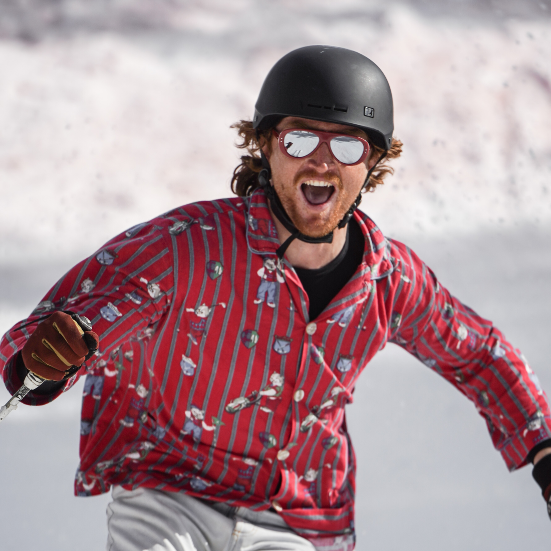 A man skiing in the snow wearing a red and gray vertically striped button-up shirt with a cartoon dog print, a black helmet, and mirrored Ski Aviators sunglasses from VALLON.