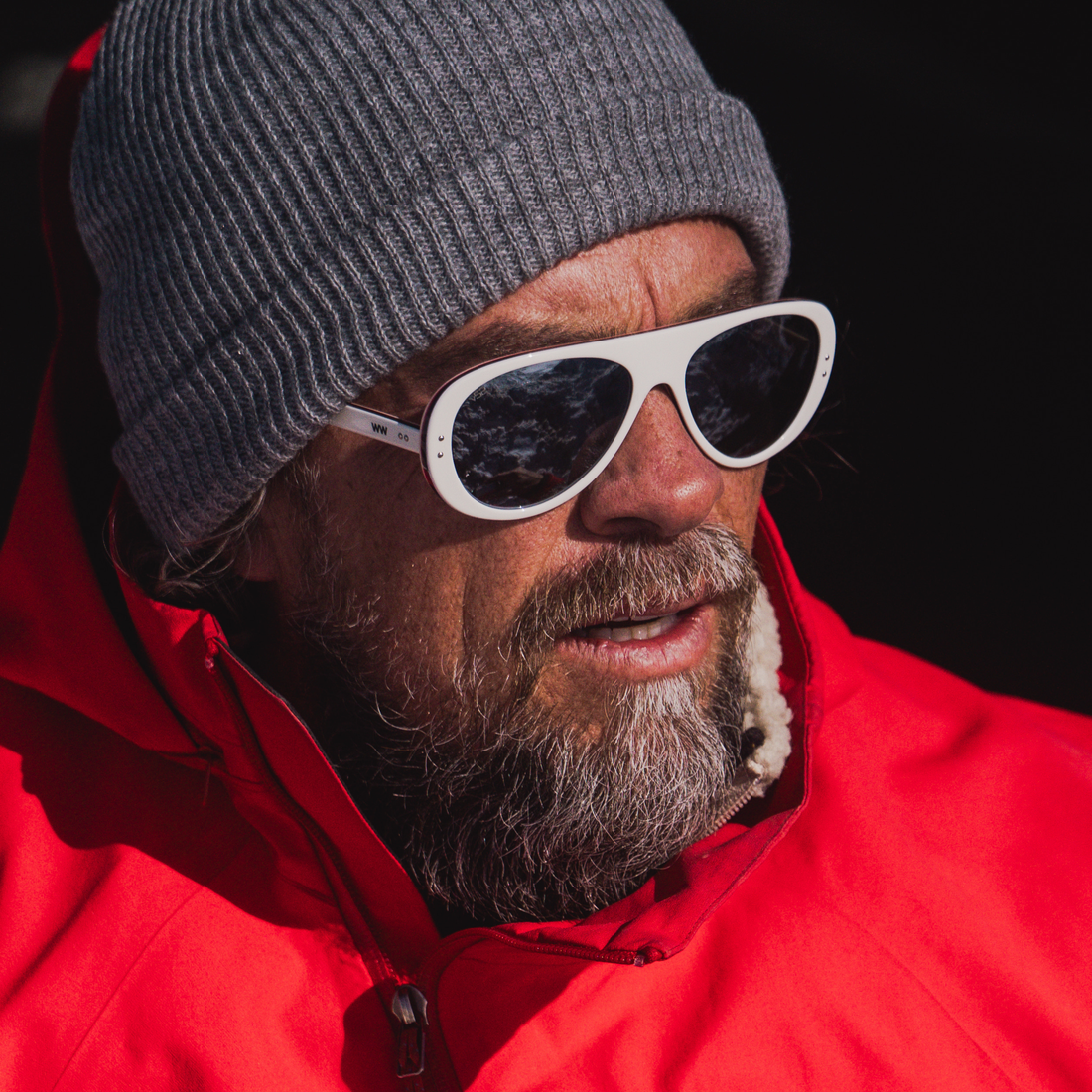 A close-up shot of a man wearing white aviator-style sunglasses  from VALLON with dark lenses, a gray ribbed knit beanie, and a bright red jacket against a dark background.