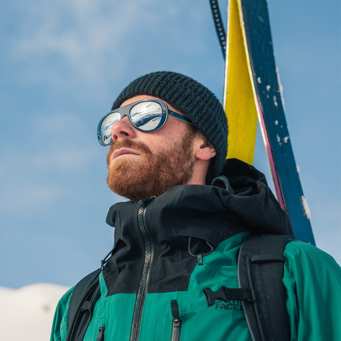 A man wearing a black beanie, Tricolor Blue Ski Aviators sunglasses from VALLON, and a green and black ski jacket with a backpack, carrying skis against a blue sky background.