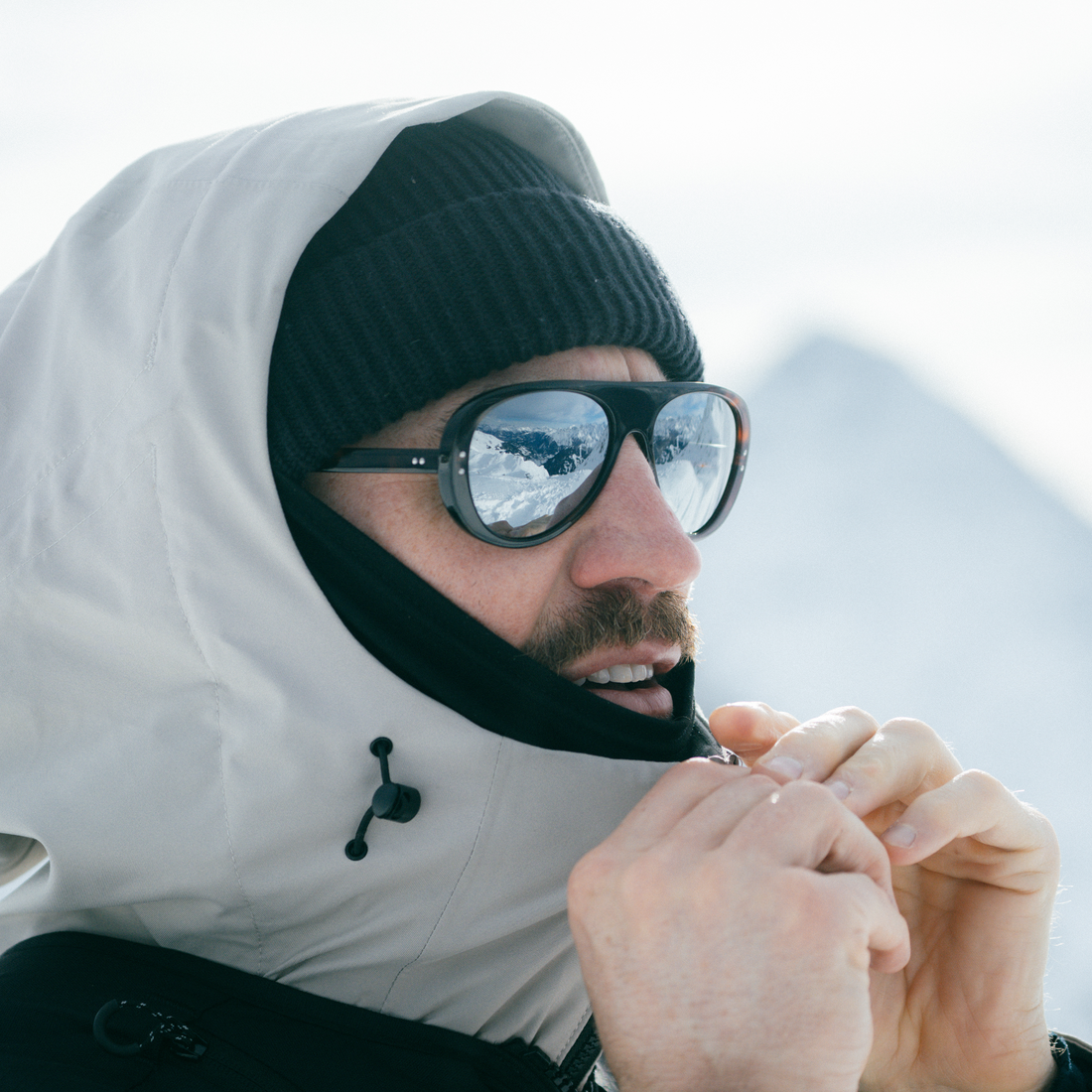 A close-up lifestyle shot of a man wearing a white hooded winter jacket with a black neck gaiter, a black knit beanie, and dark tortoise VALLON Ski Aviators sunglasses. The background is a bright, snowy mountain landscape.
