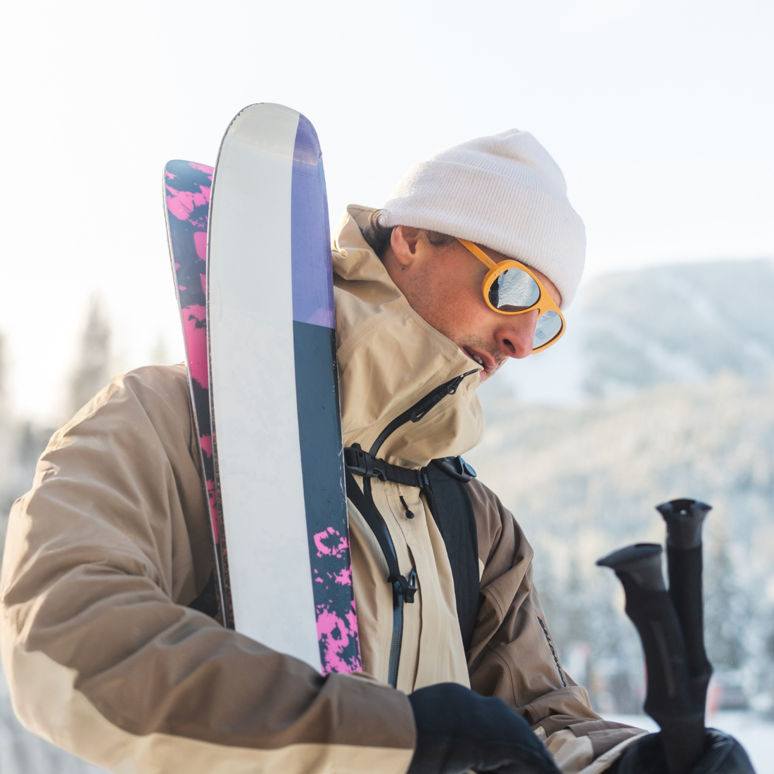 A man in a beige ski jacket, white beanie, and yellow-framed Ski Aviators from VALLON carries a pair of skis with pink and purple graphics and black ski poles in a snowy outdoor setting.