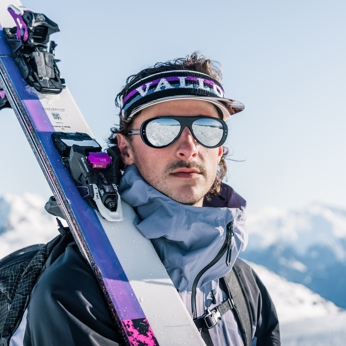 A close-up portrait of a man in ski gear on a snowy mountain, wearing a VALLON Vizor, tricolor blue VALLON Ski Aviators, a gray ski jacket, and carrying skis over his shoulder.