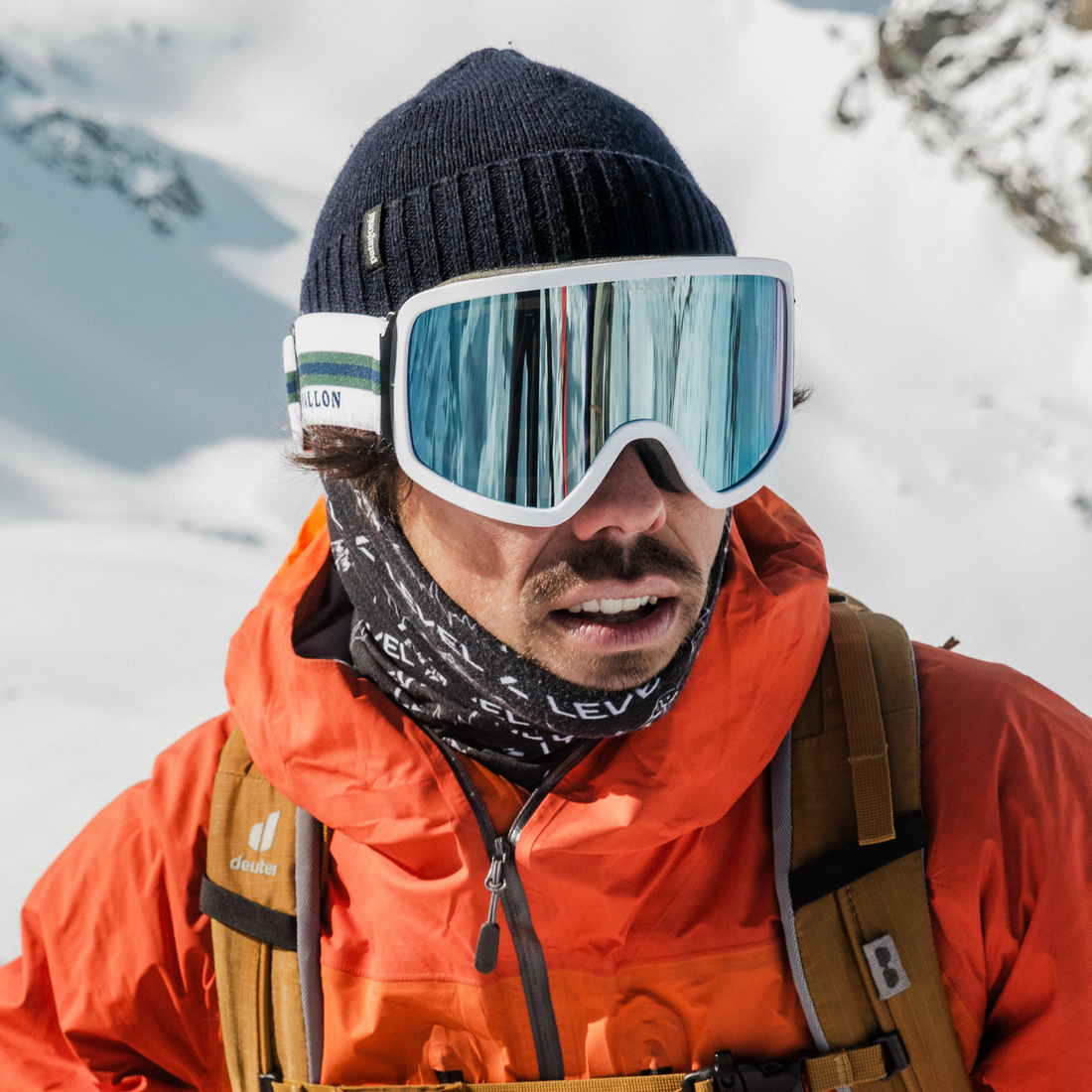 Male skier wears VALLON Stairways white goggles with blue reflective lenses, with a navy blue beanie, orange ski jacket, dark yellow backpack and a black neck buff