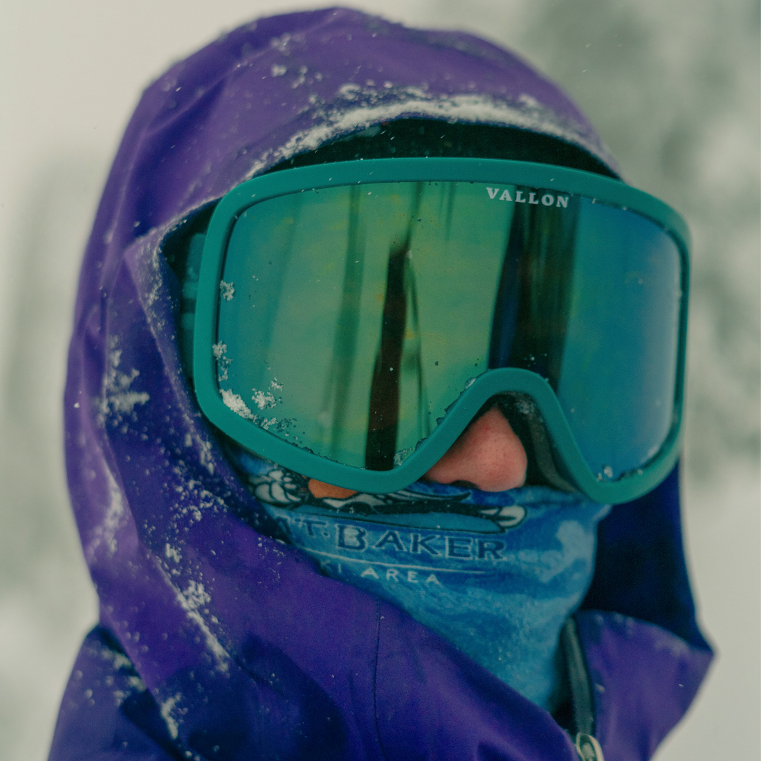 Person looks directly at the camera in fresh snowfall wearing a bright purple ski jacket with the hood up and Teal-Green Stairways ski goggles from VALLON, paired with a blue neck buff that comes up to their nose.