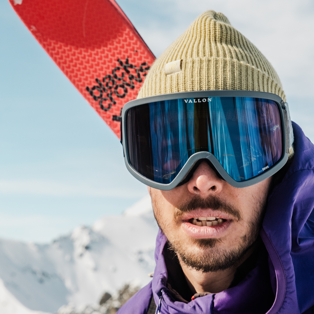 Man wears VALLON Stairways ski goggles with a blue frame and dark blue mirror lenses, along with a beige beanie and purple softshell jacket while carrying red Blackcrows skis on his back. Snowcapped mountains are blurred in the background.