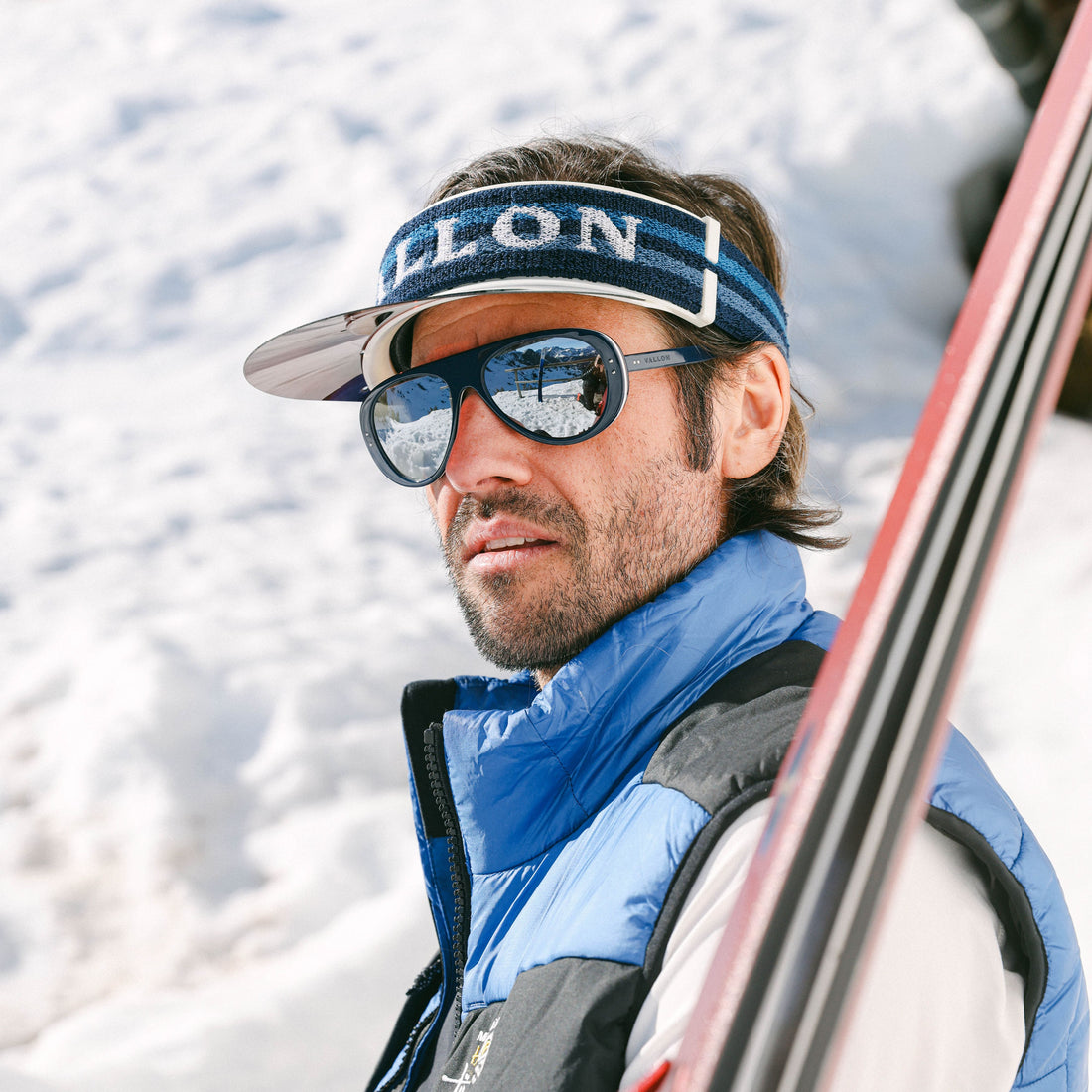 Man wears VALLON Vizor in blue with ski aviators and a pair of skis outside a mountain lodge