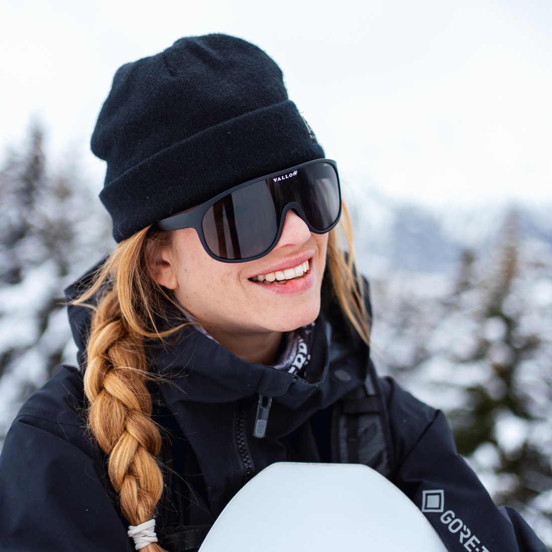 A woman outdoors in a snowy setting wearing a black knit beanie, black Watchtowers ski sunglasses with the VALLON brand visible on the bridge, and a black winter jacket.