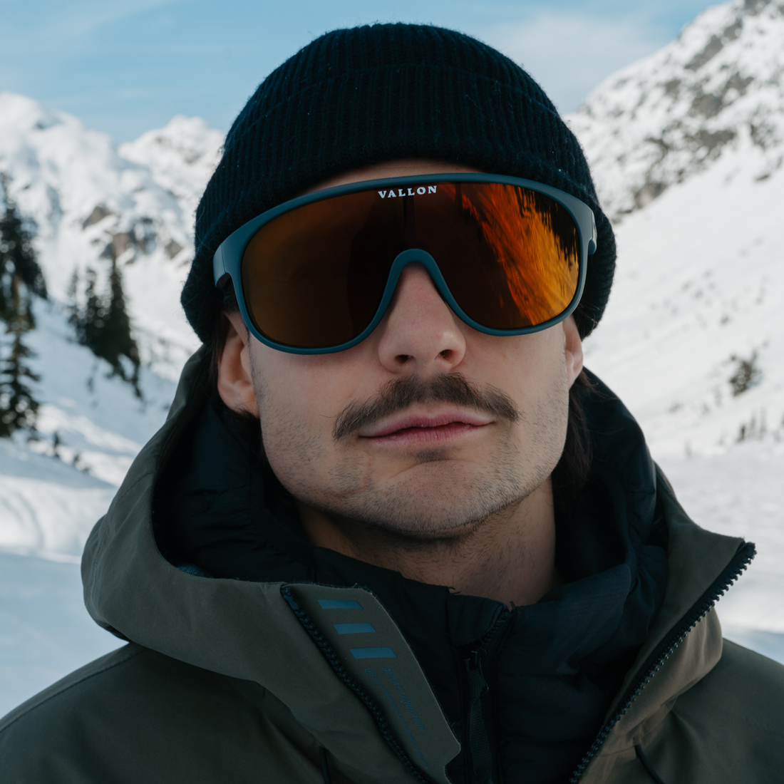 Close up of man wearing dark green ski jacket a black beanie and VALLON Dark Teal Copper Watchtowers sunglasses in a snowy alpine setting
