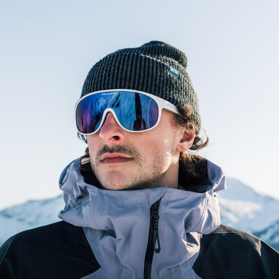 A skier looking to one side in an snowy mountain setting wearing a purple ski jacket, dark grey wool beanie and Silver-Blue VALLON Watchtowers sunglasses with blue mirror lenses and a bright silver frame