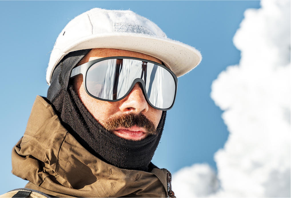 A close-up of a man wearing VALLON Watchtowers ski goggles with a black frame and full silver mirrored lenses, a black neck gaiter, a brown jacket, and a white cap against a blue sky with clouds.