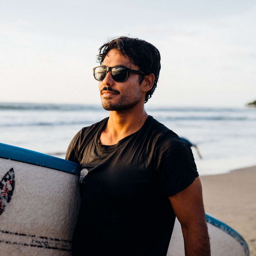 Man wears Wilburys Rx sunglasses from VALLON while carrying a surfboard at the beach